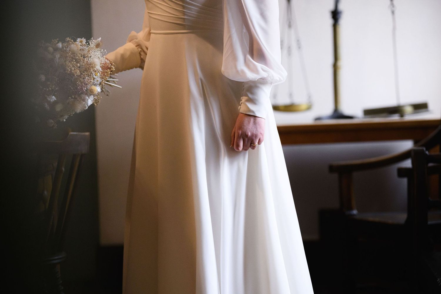 A flowing white dress catches light near a window, creating an ethereal bridal portrait with soft natural lighting.