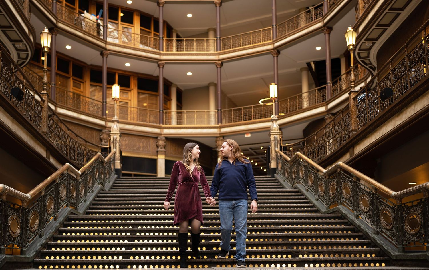 Couple holding hands on grand ornate staircase inside historic building with elegant balconies.