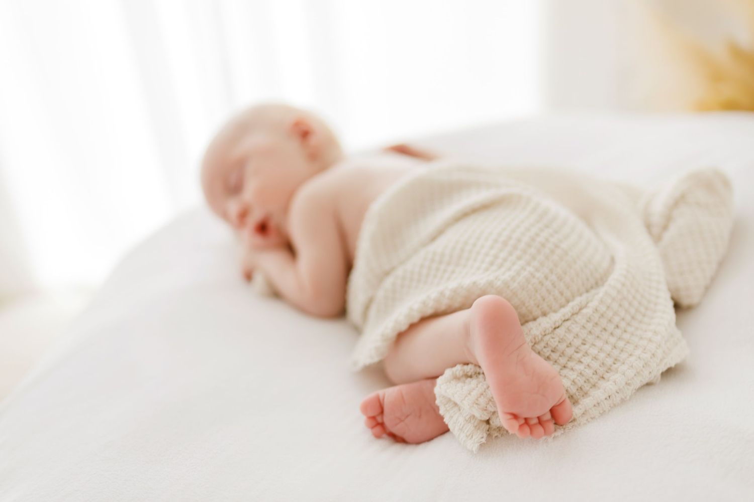 Newborn baby sleeping peacefully on white surface wearing cream knit outfit with tiny feet visible.