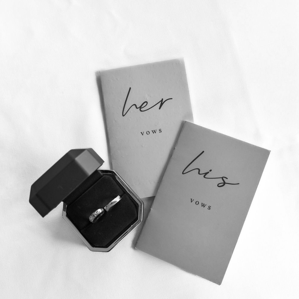 His and her vow booklets in gray with a diamond ring in black octagonal box on white background.