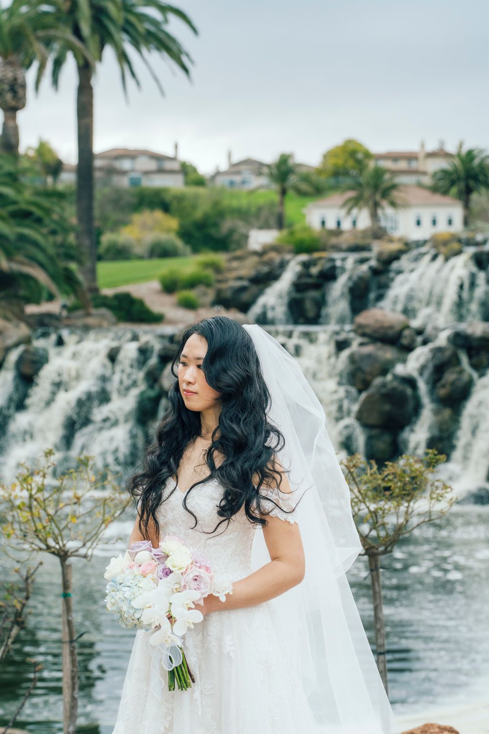 Beautiful Asian bride in white lace gown holds bouquet before waterfall backdrop on wedding day.
