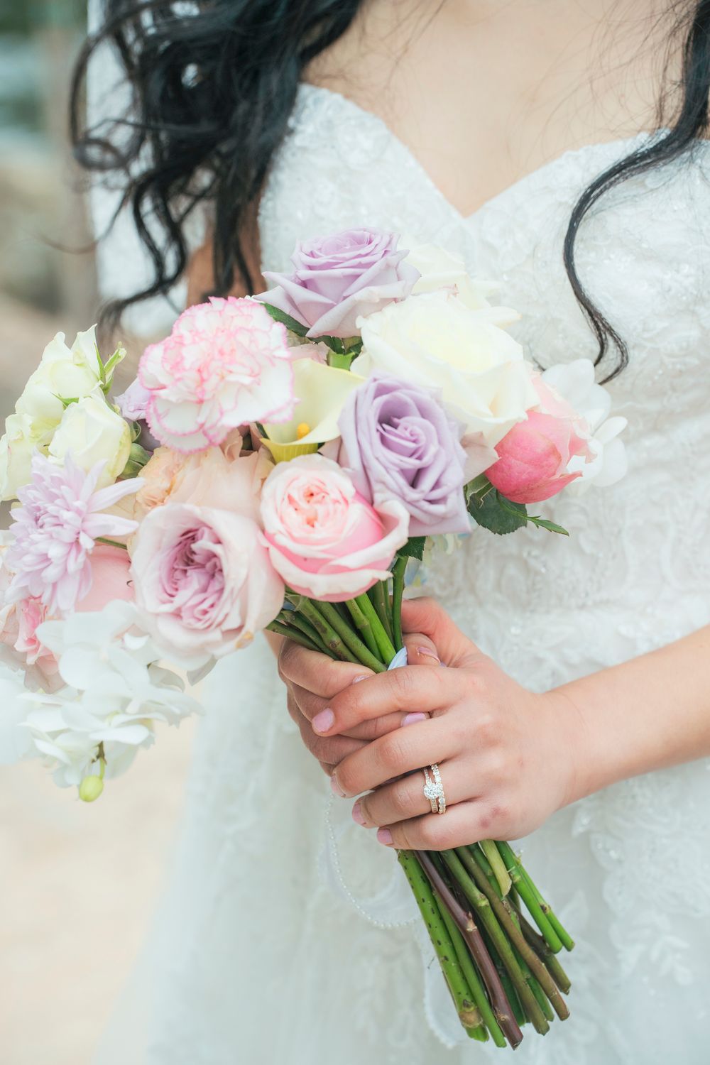 Bride in white lace gown holding pastel bouquet of pink and purple roses with diamond ring on finger.