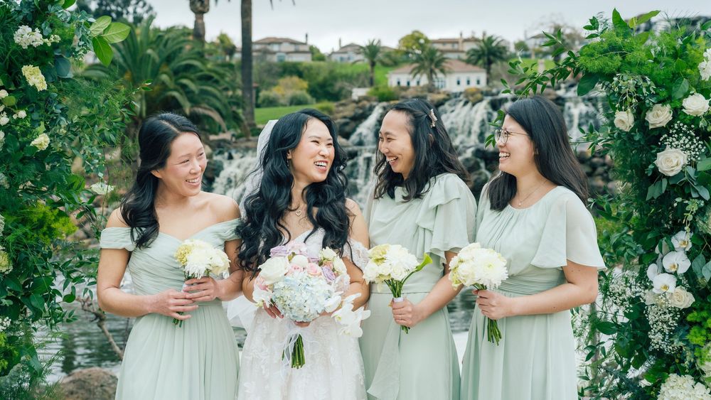 Bride and three bridesmaids in sage green dresses laughing together outdoors with floral arch and waterfall backdrop.