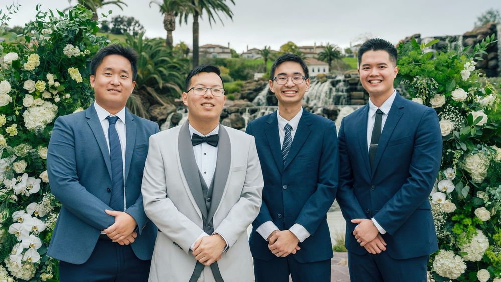 Four men in formal suits smiling together outdoors at a wedding ceremony with white floral arrangements.