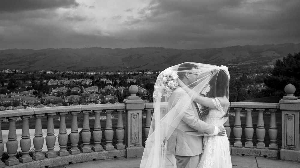 Black and white wedding photo of bride and groom kissing under veil on balcony with mountain views.