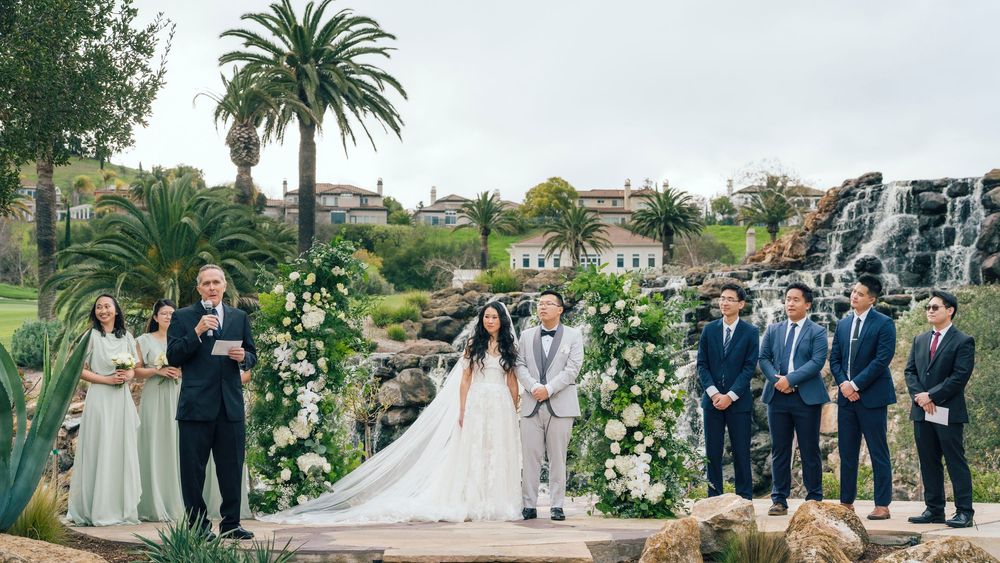 Elegant outdoor wedding ceremony with bride and groom at floral arch, waterfall backdrop, groomsmen, and lush palm trees.