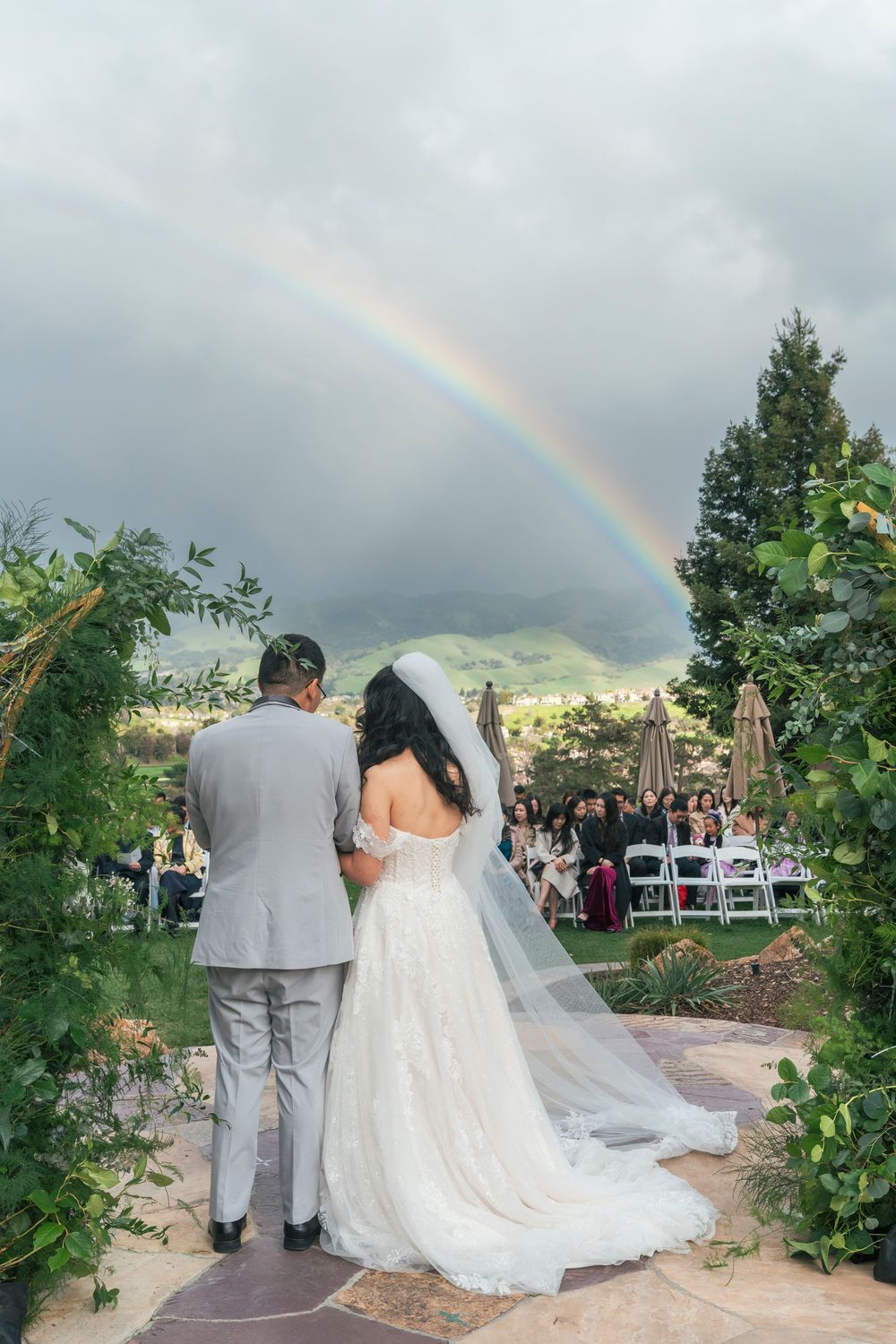 Bride and groom stand before a stunning rainbow during outdoor wedding ceremony with mountain views and seated guests.