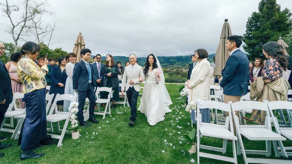 Bride walks down the aisle at outdoor wedding ceremony on green lawn with white chairs and guests.