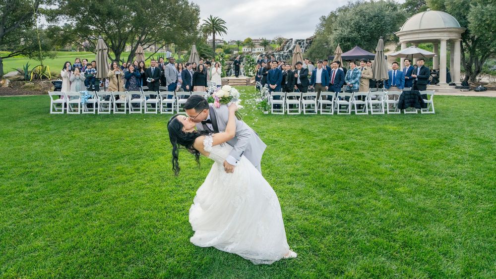 Couple shares romantic dip kiss on green lawn at outdoor wedding ceremony with guests seated in white chairs.