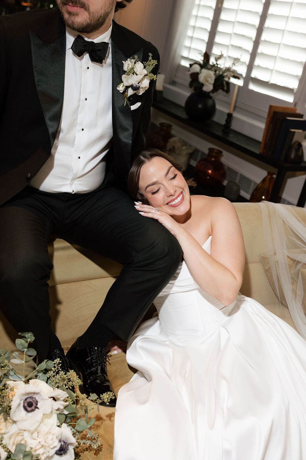Bride in white gown laughs joyfully leaning on groom in black tuxedo with floral boutonniere at indoor wedding.