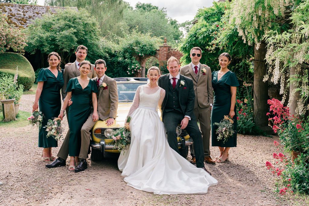 Wedding party poses by vintage gold car, bride in white gown, bridesmaids in green dresses, groomsmen in tan suits.