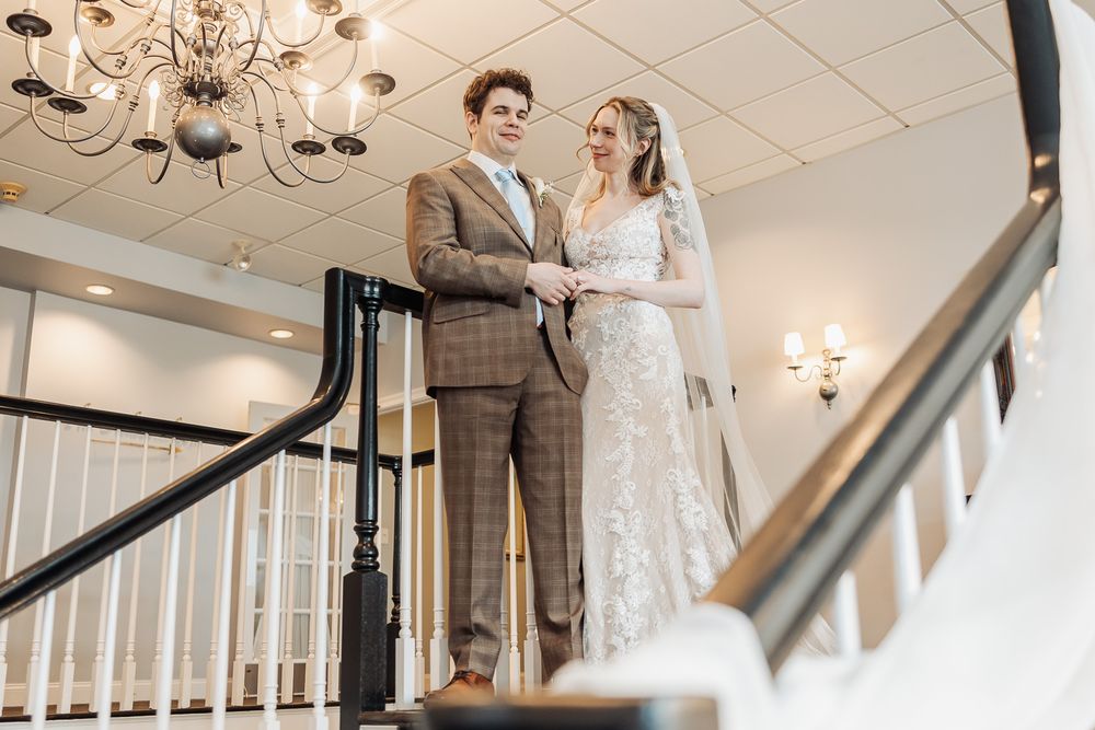 Bride and groom stand together on elegant staircase, she in lace gown with veil, he in plaid suit, beneath chandelier.