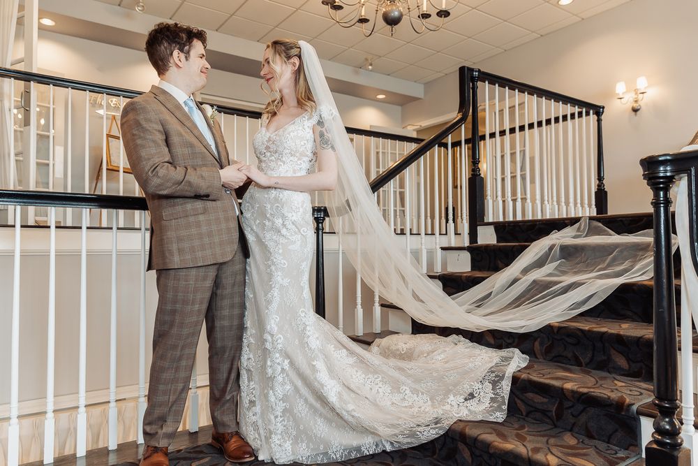 Bride and groom holding hands on elegant staircase, bride wearing lace wedding gown with cathedral veil.