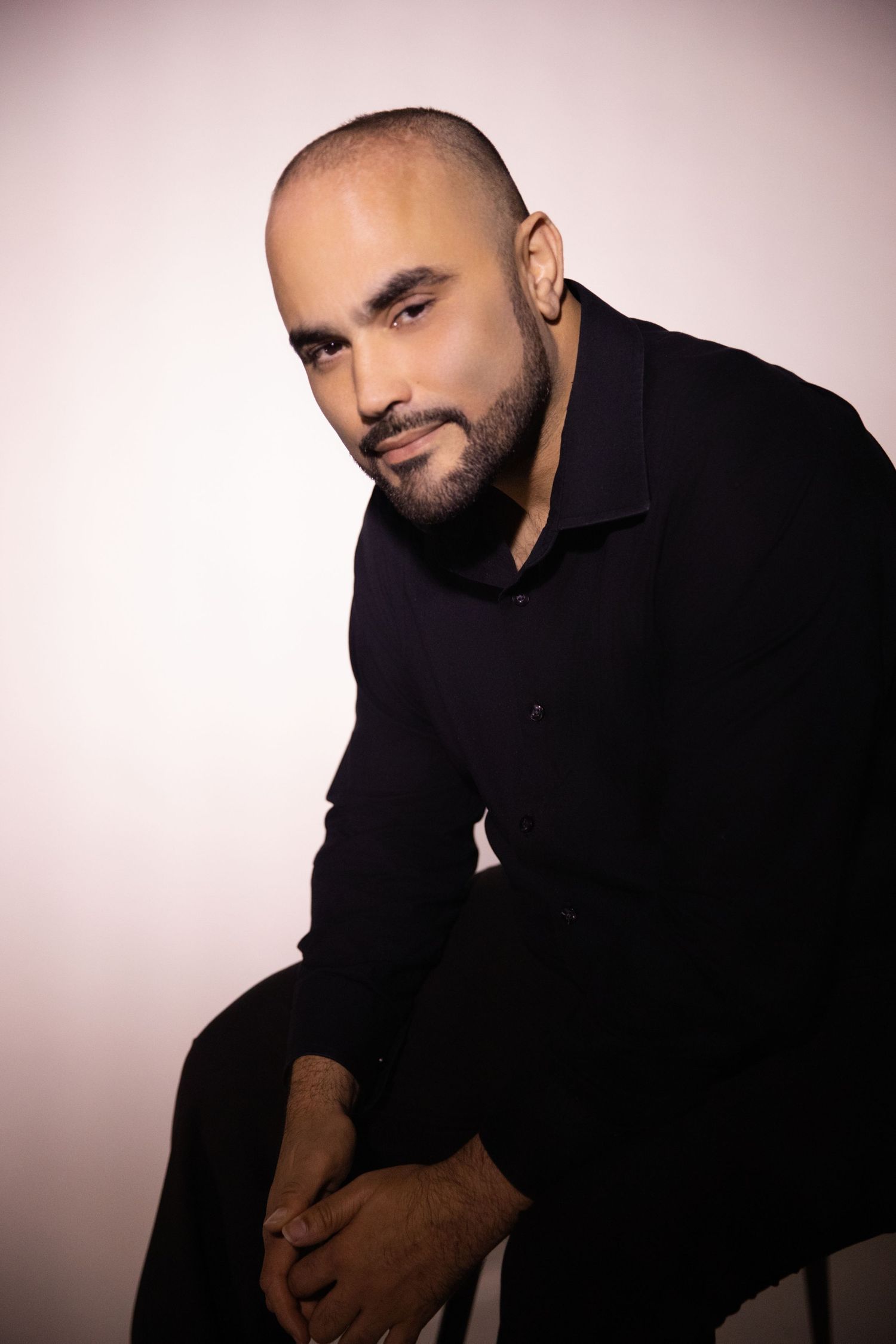 A confident bald man with a beard wearing a black shirt, sitting and posing against a light background.