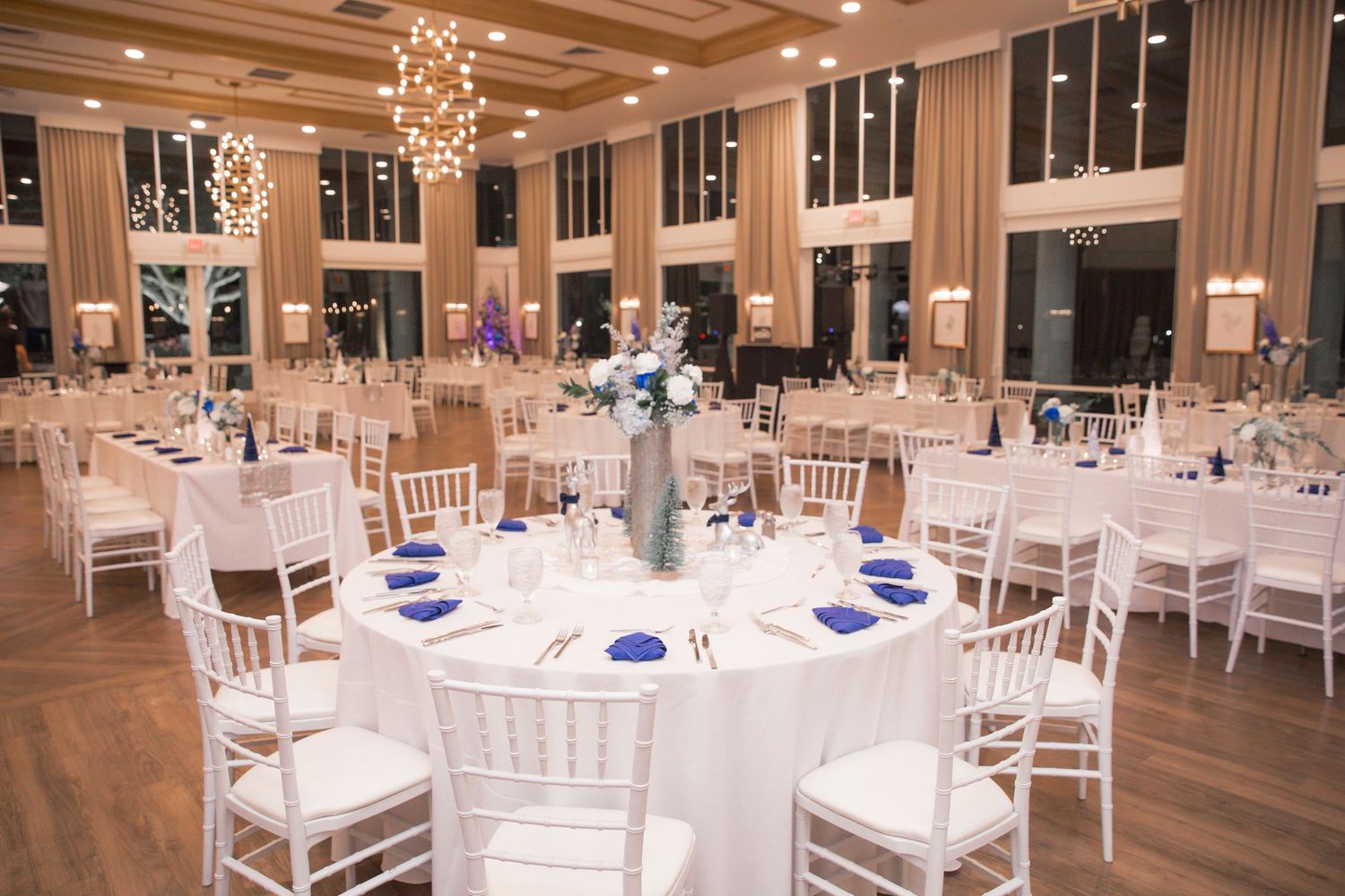 Elegant wedding reception hall with white chiavari chairs, round tables with blue napkins, and warm chandelier lighting.