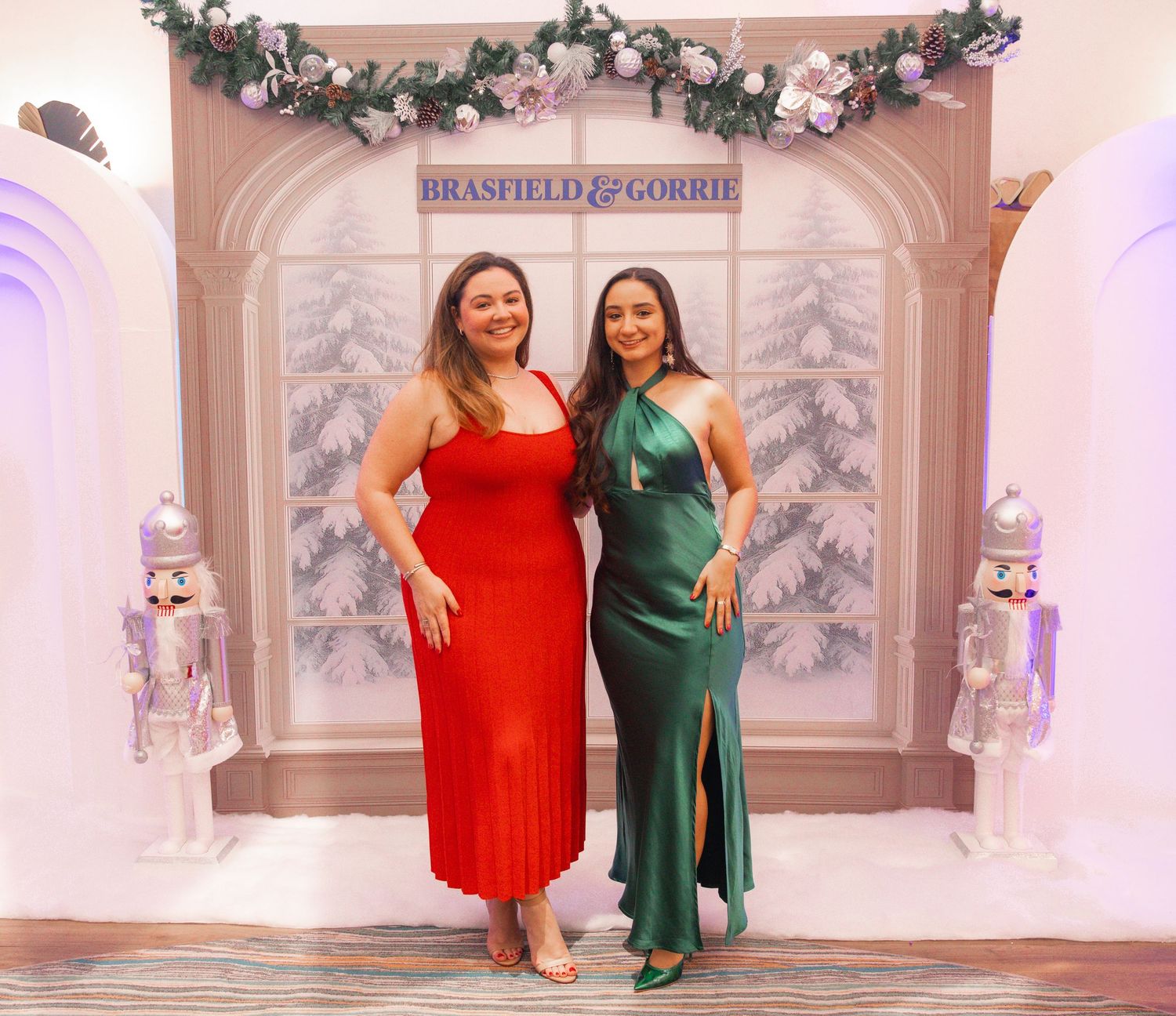 Two women in elegant red and green dresses pose together at a festive Brasfield & Gorrie holiday party backdrop.