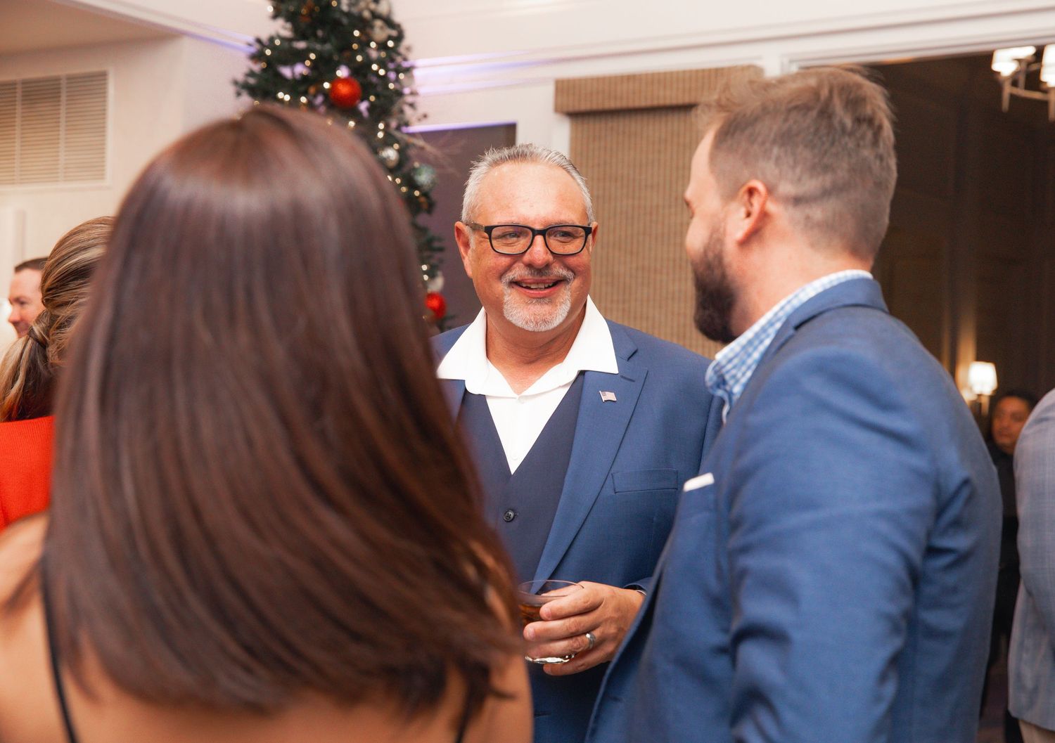 Three people socializing at a festive Christmas party near a decorated tree in an elegant venue.
