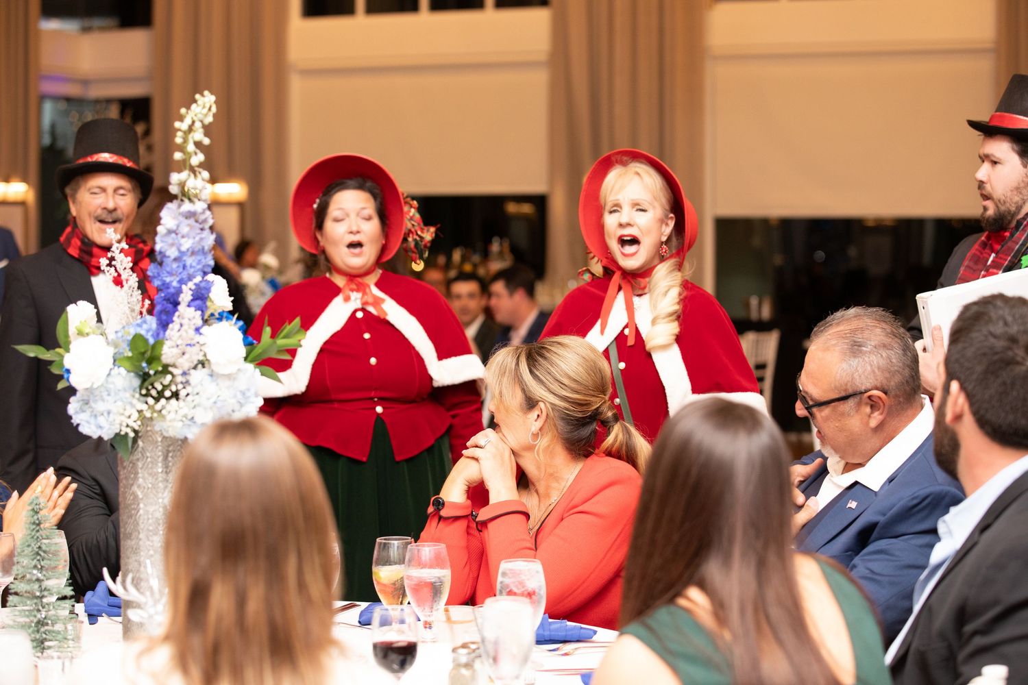 Christmas carolers in red capes perform for guests at an elegant holiday dinner event.