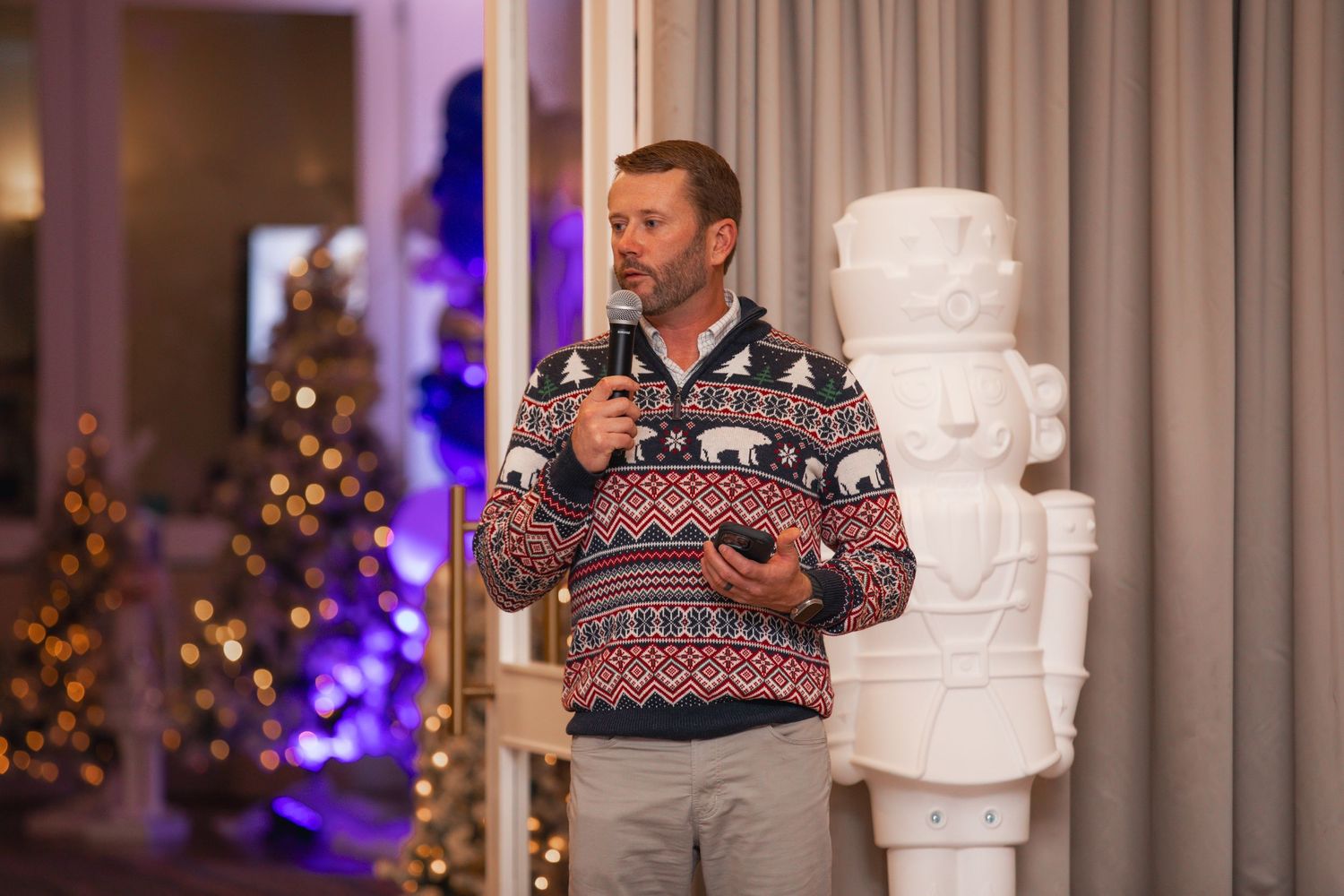 Man in festive holiday sweater speaking into microphone at Christmas party with decorated tree and nutcracker decor.