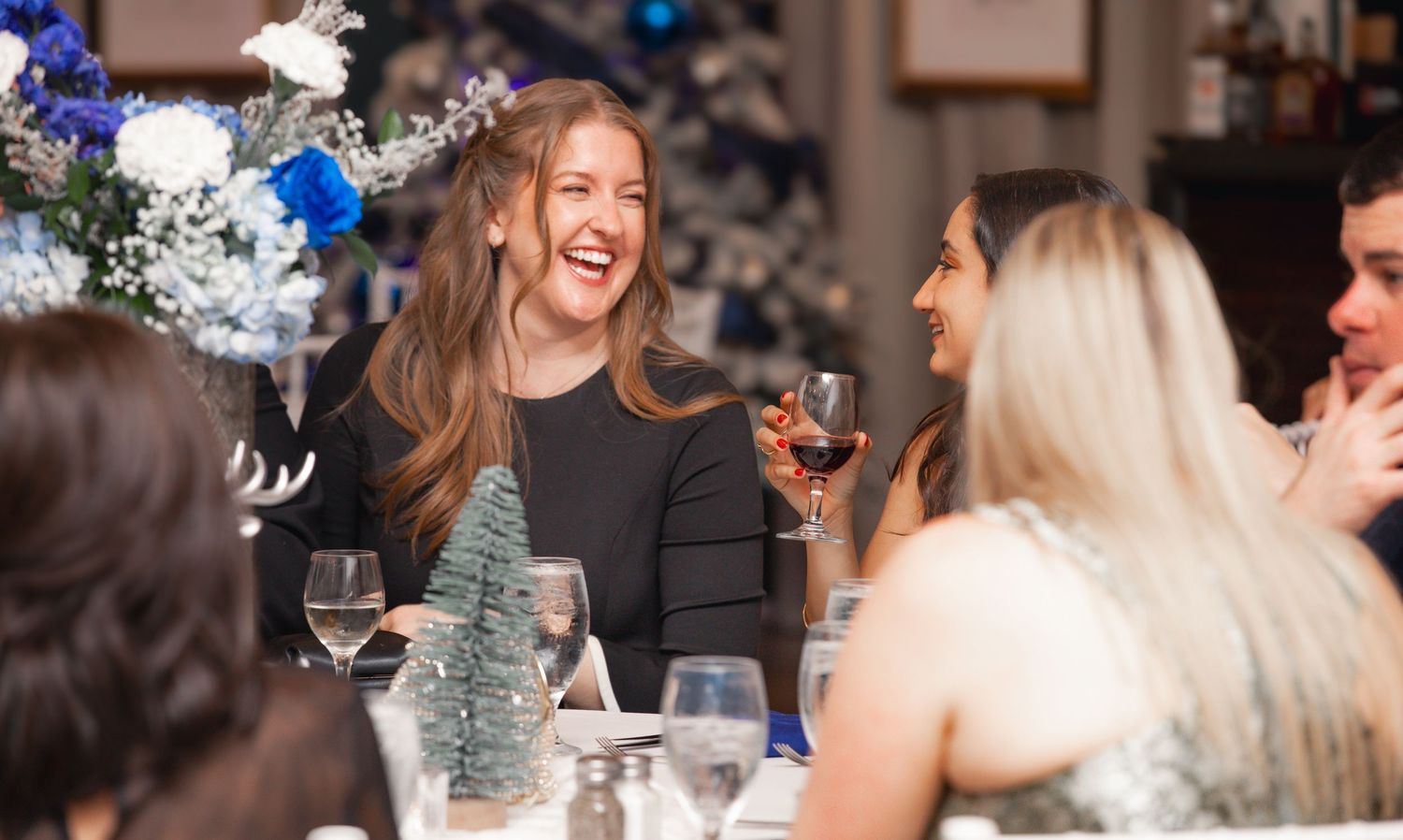 Women laughing and enjoying wine at a festive holiday dinner party with blue and white floral decorations.