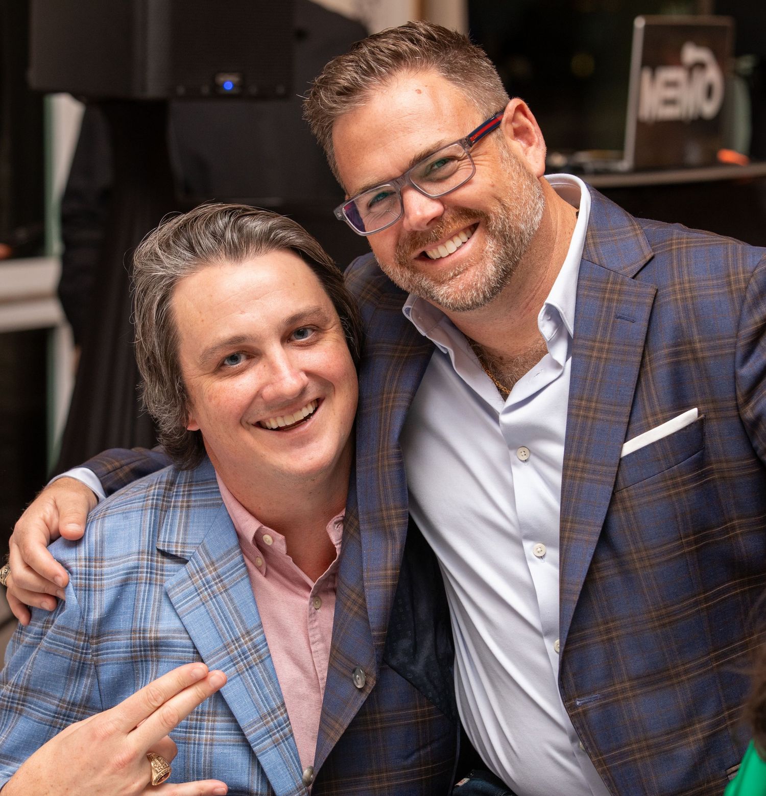 Two smiling men in blazers pose together with arms around each other at a social event.