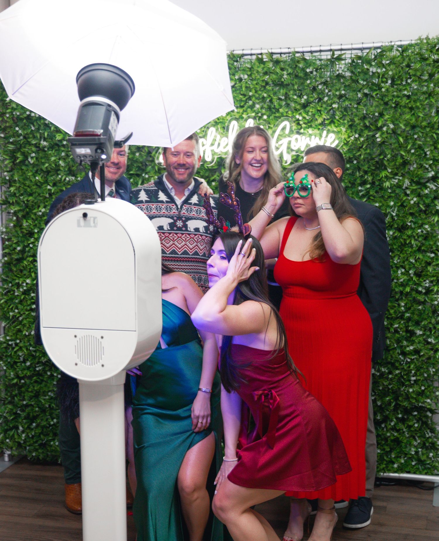 Group of friends posing at a photo booth with green hedge backdrop, wearing festive attire and fun masks.