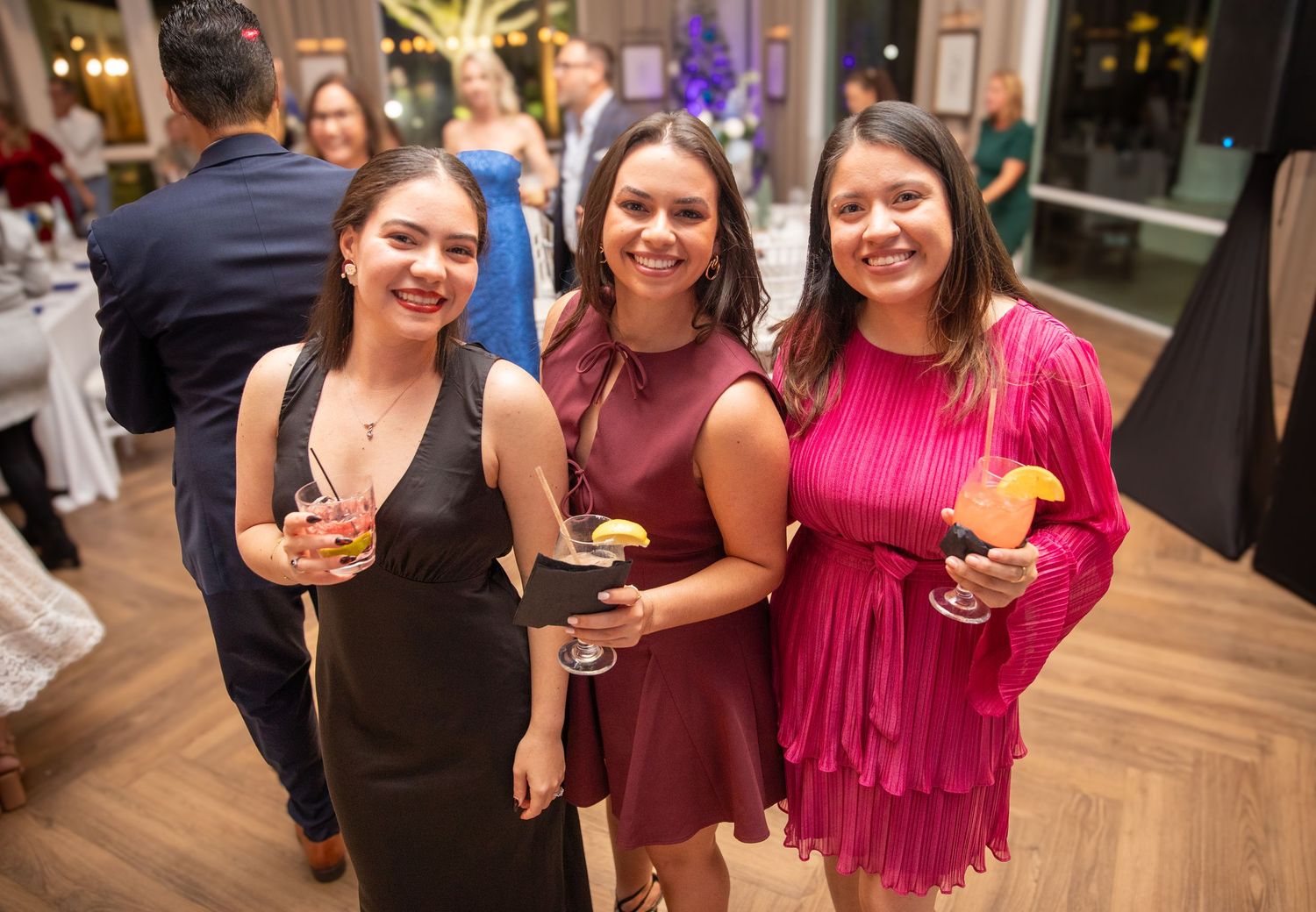Three women in elegant dresses smile and hold cocktails at a formal evening party event.