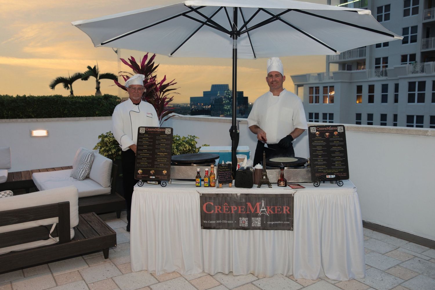 Two chefs in white uniforms stand behind a CrepeMaker station under an umbrella on a rooftop terrace at sunset.