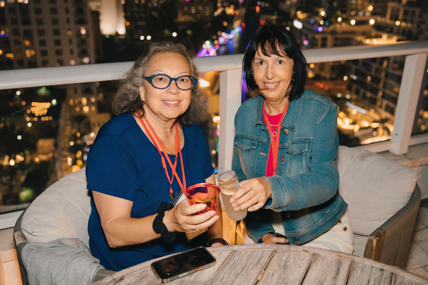 Two women smiling and holding drinks on a rooftop bar at night with city lights in the background.