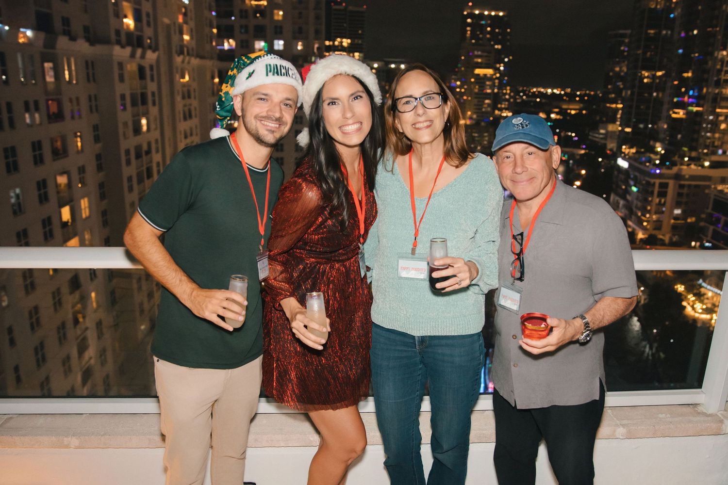 Four friends pose together at a festive rooftop holiday party with city skyline views at night.
