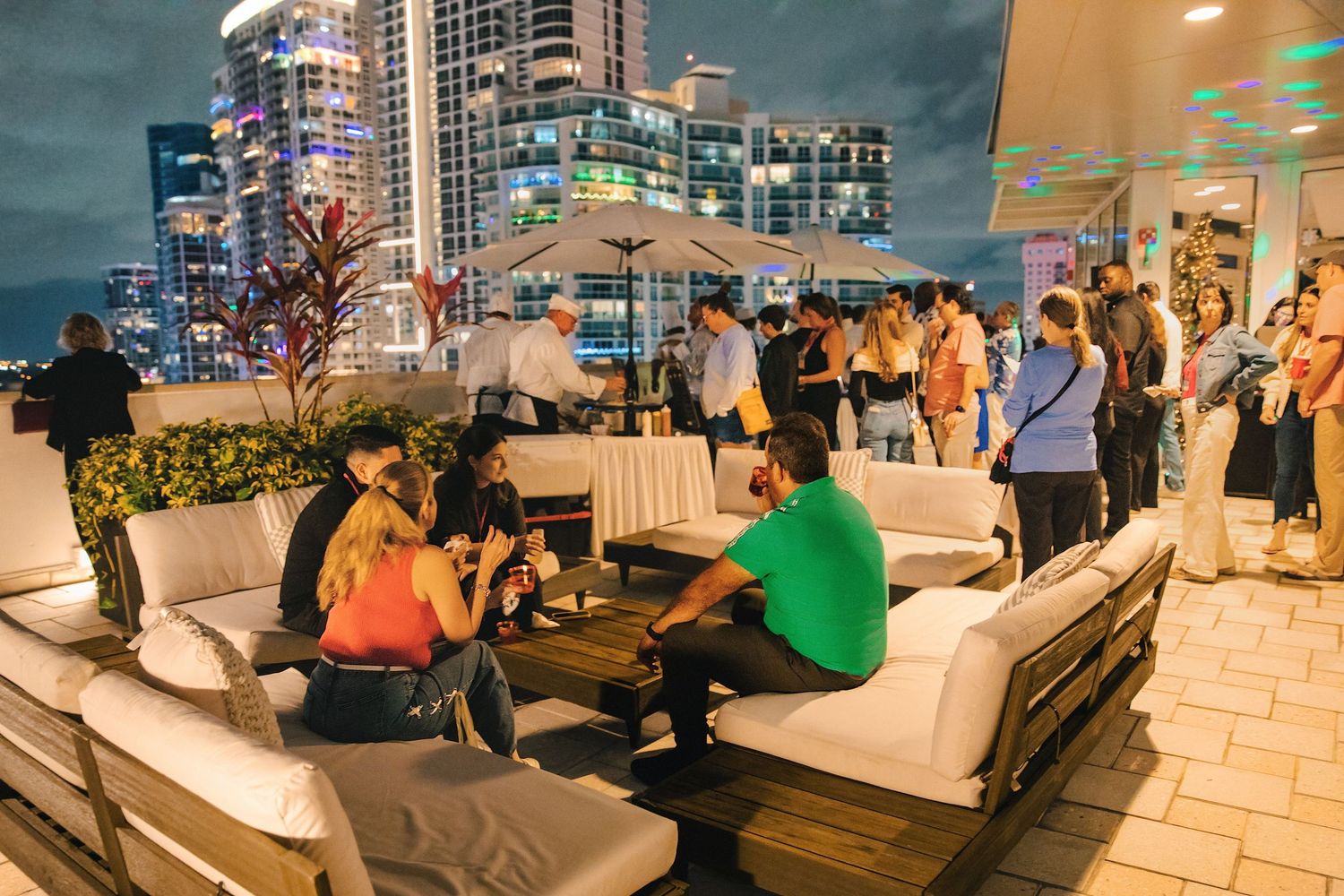 Rooftop bar party at night with city skyline, guests lounging on white sofas and mingling under colorful lights.