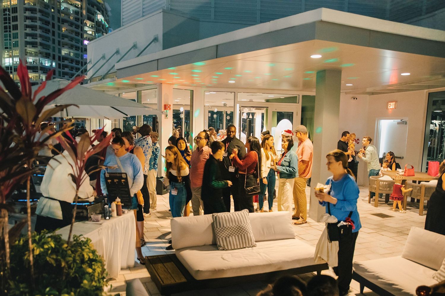 Guests mingle at an upscale rooftop event with city skyline views, white lounge furniture, and festive lighting at night.