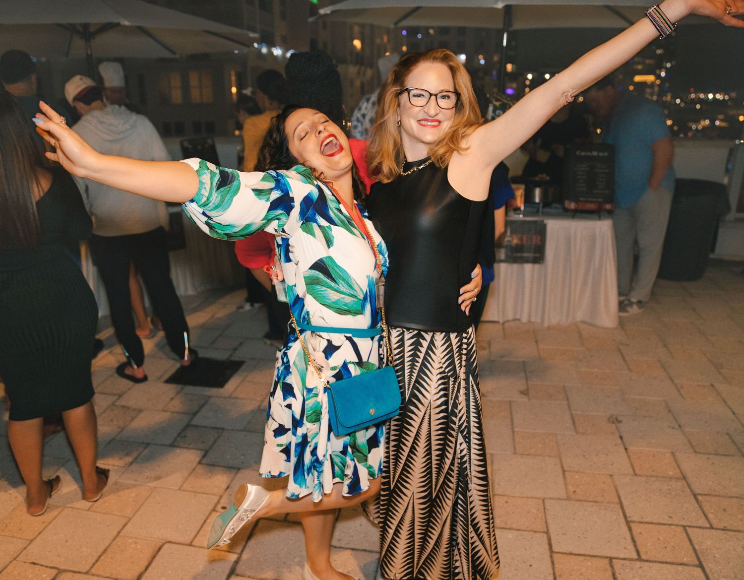 Two women celebrating at an outdoor evening party, arms raised joyfully, wearing colorful and elegant dresses.