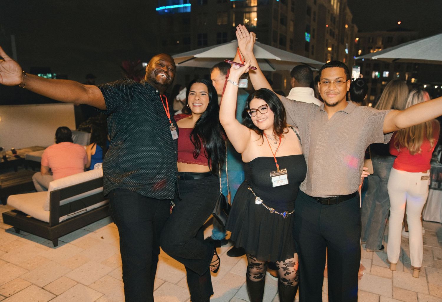 Four friends celebrate at an outdoor evening party, raising hands together and smiling for the camera.