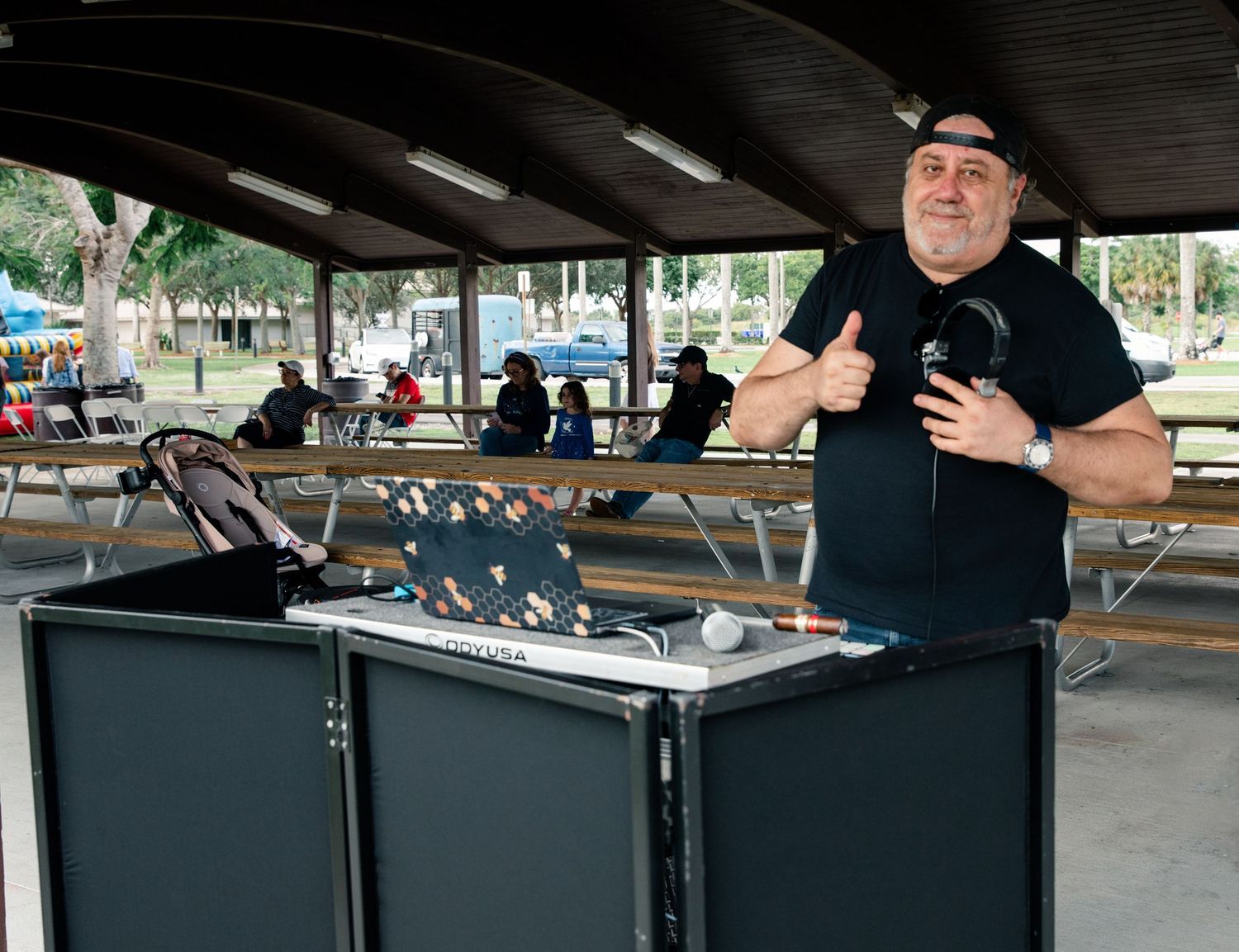 A DJ giving thumbs up behind his equipment setup at an outdoor covered pavilion event.