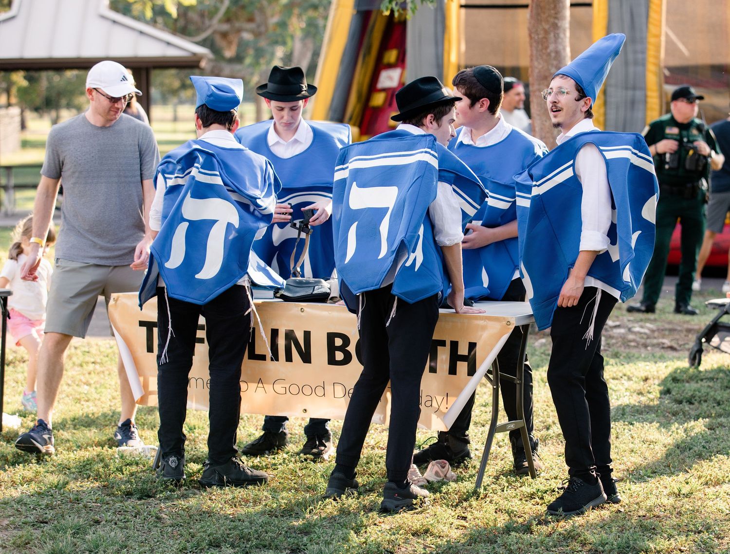 Group of young men in blue dreidel costumes gather around a Tefillin Booth sign at an outdoor Jewish community event.