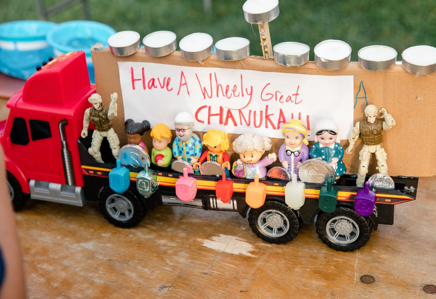 A toy truck decorated with action figures and a Chanukah sign reading Have A Wheely Great Chanukah with a menorah backdrop.