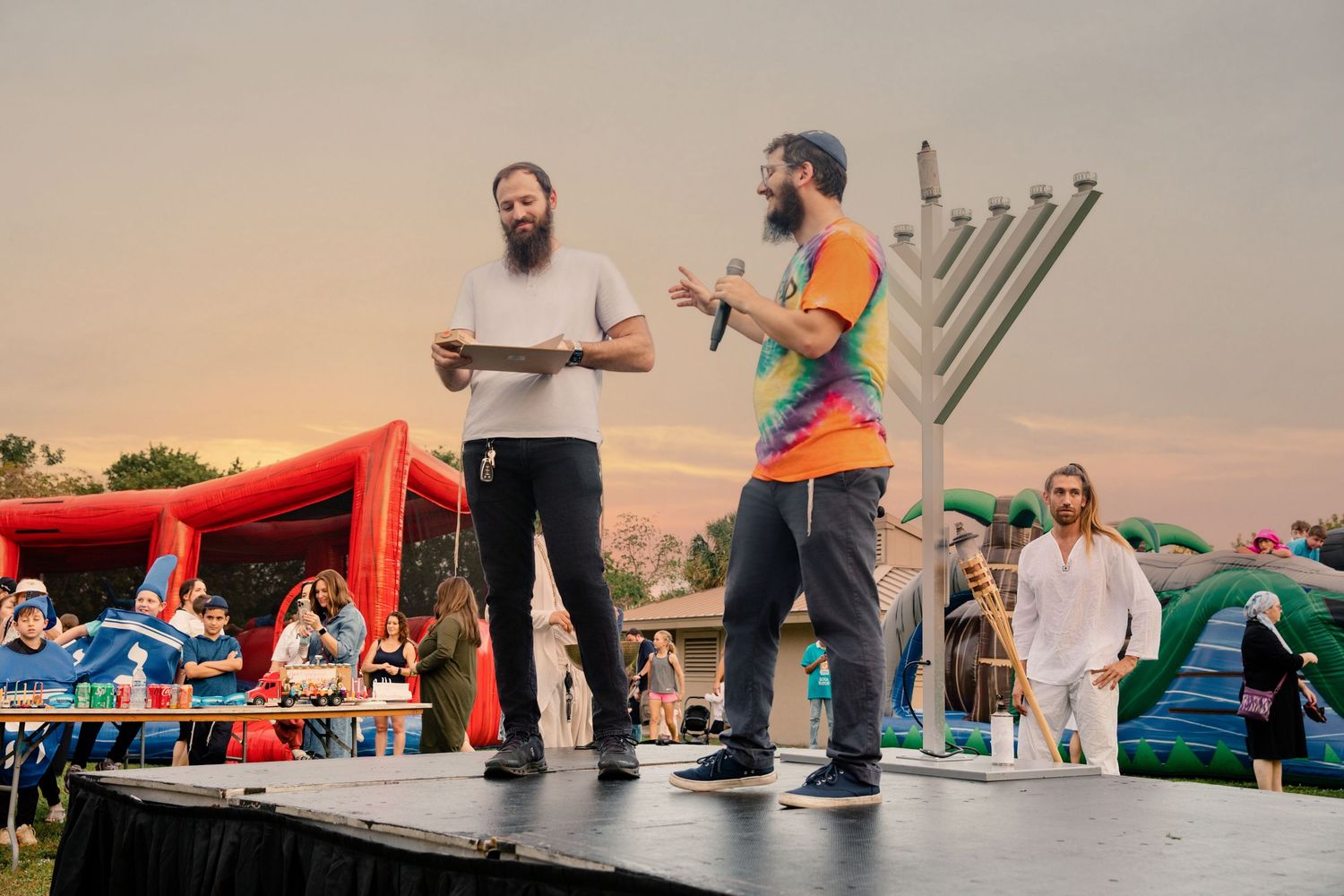 Two bearded men on outdoor stage at Jewish festival with menorah backdrop at sunset.