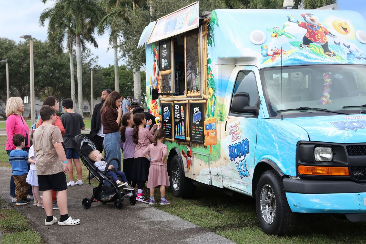 Families and children line up at a colorful snow cone food truck outdoors in a sunny Florida park setting.
