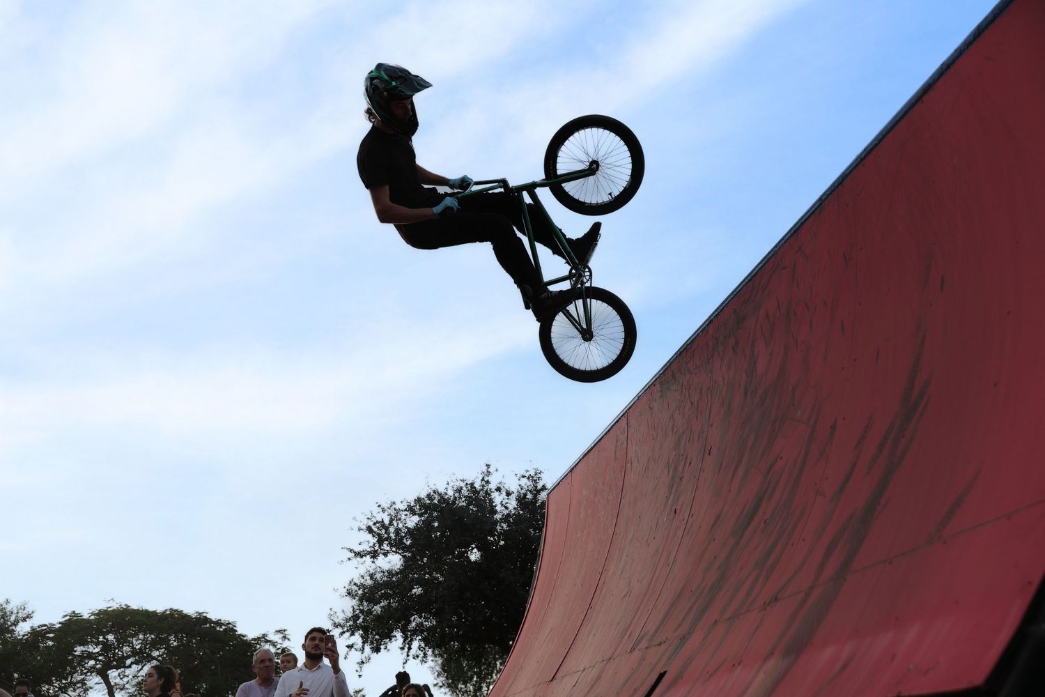 BMX rider performs a high aerial trick off a red half-pipe ramp at an outdoor skate park under a blue sky.