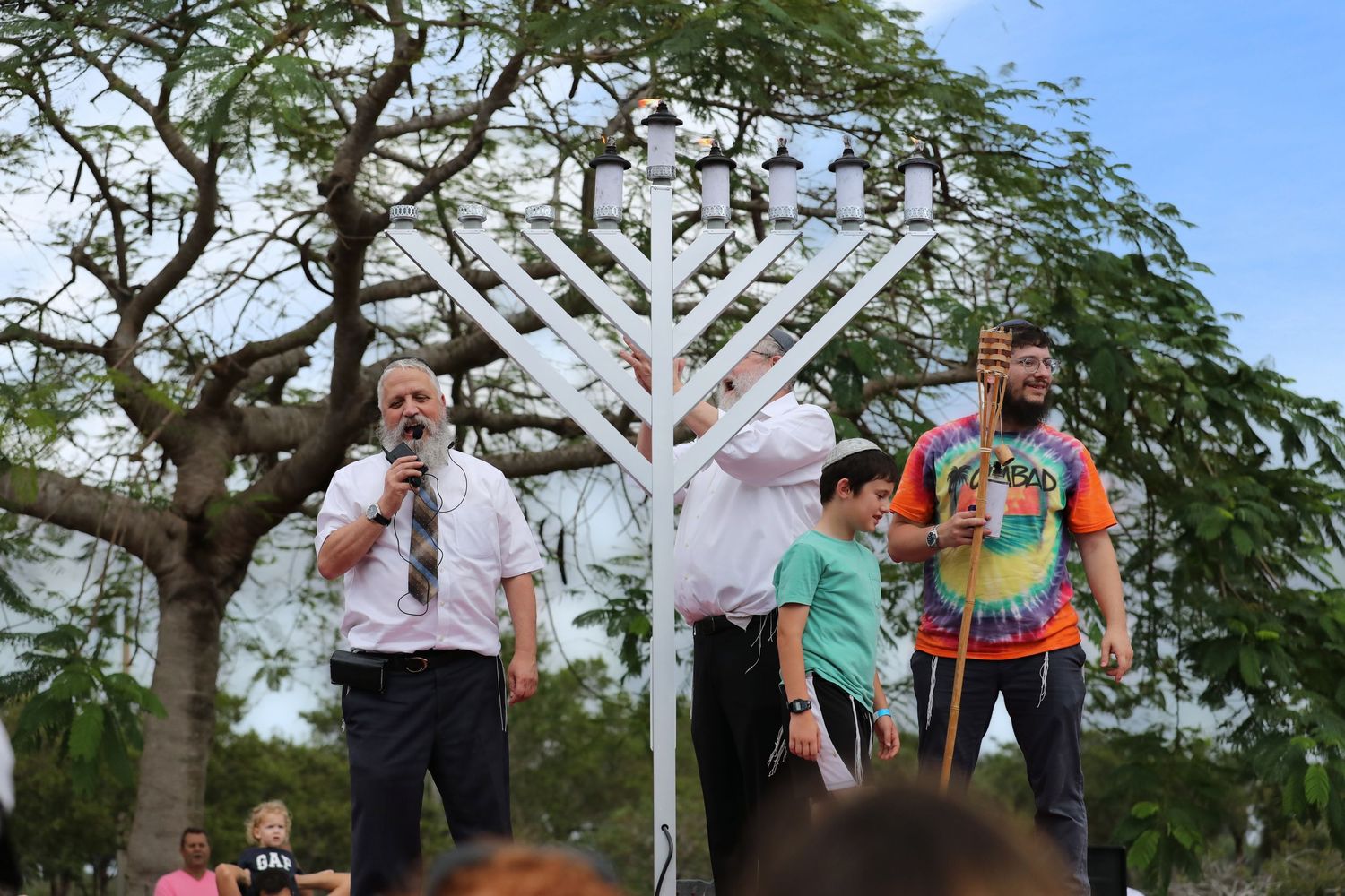 Men speaking at a Hanukkah menorah lighting ceremony outdoors in a park setting.