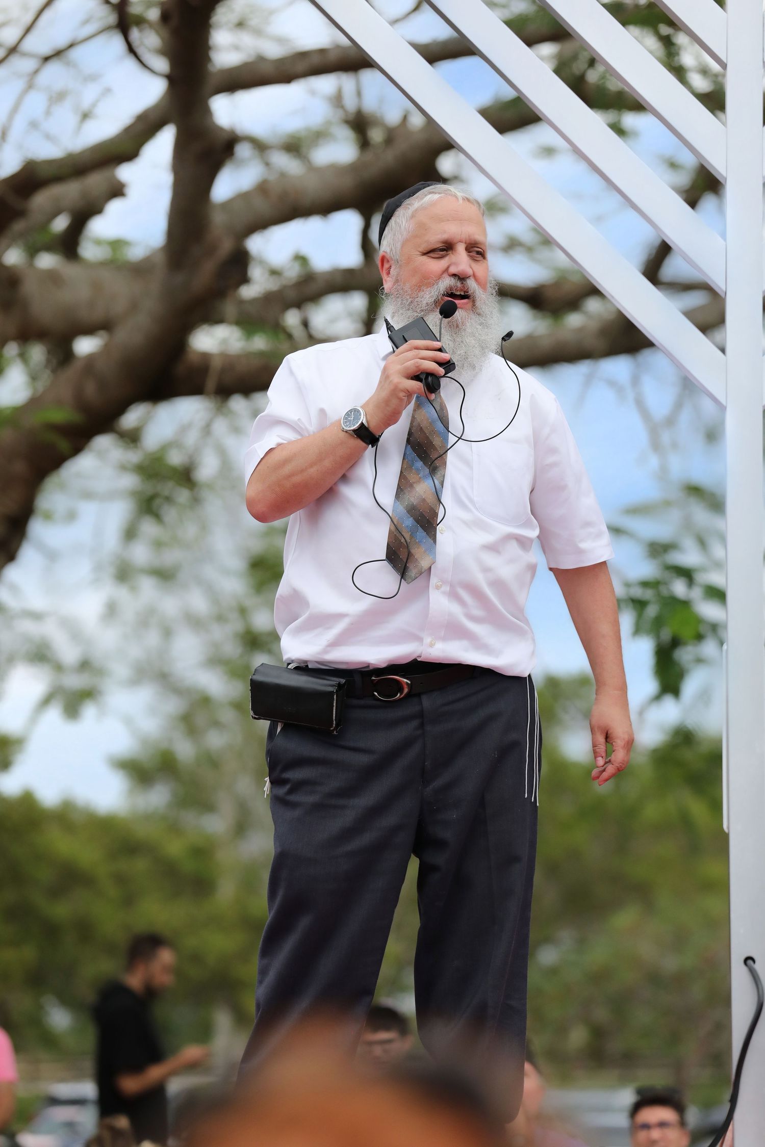 Bearded man in white shirt and tie speaks into microphone outdoors at a public event under white structure.