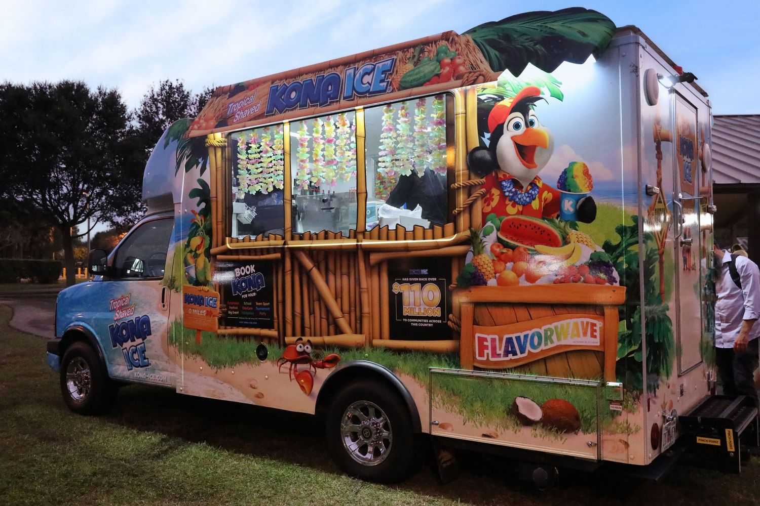 Colorful Kona Ice shaved ice truck with tropical decorations and mascot parked outdoors at dusk.