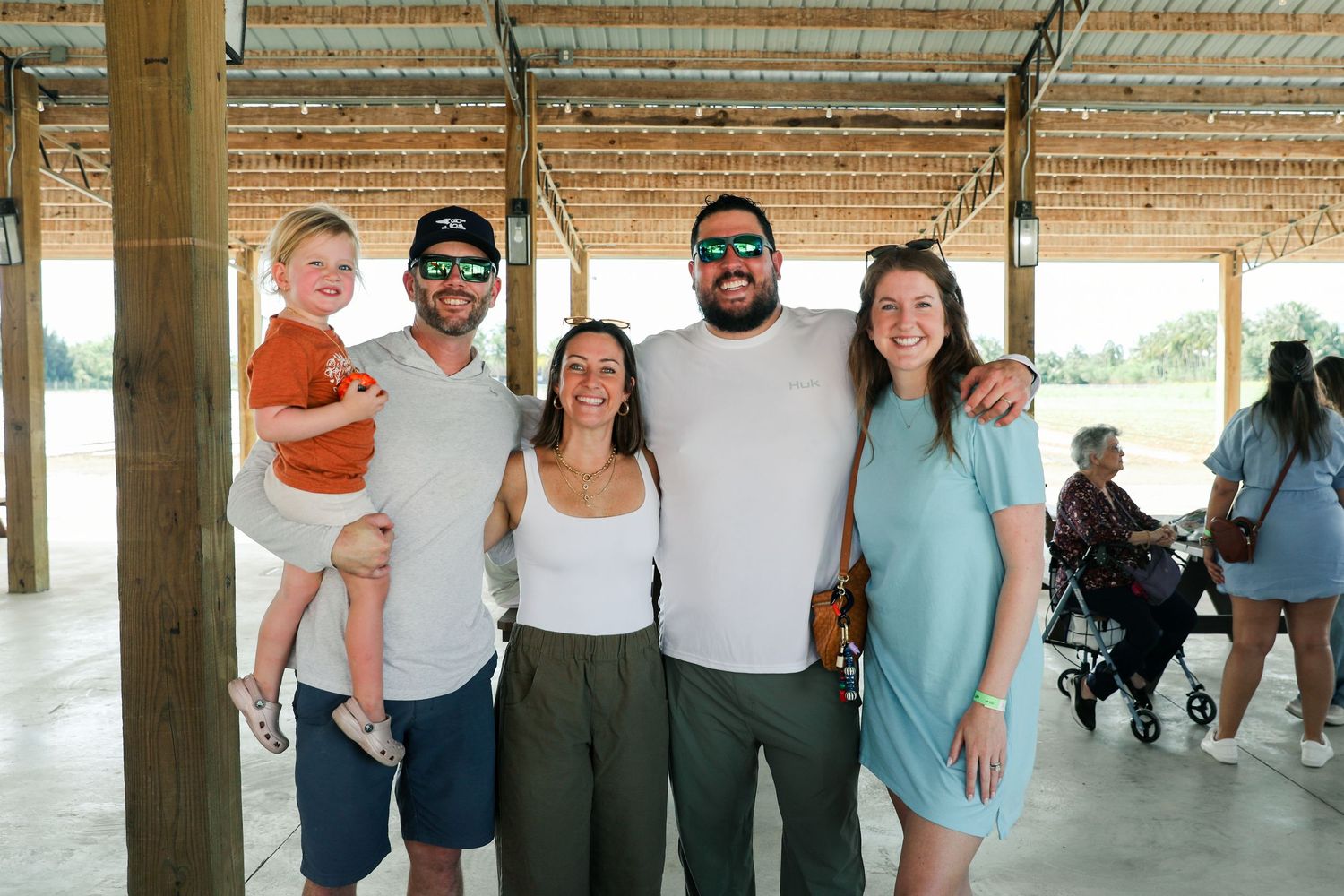 Group of five adults and a toddler smiling together under an open-air pavilion at an outdoor event.