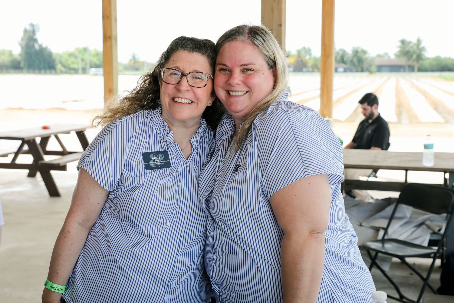 Two women in matching blue striped shirts smile together at an outdoor farm event under a covered pavilion.