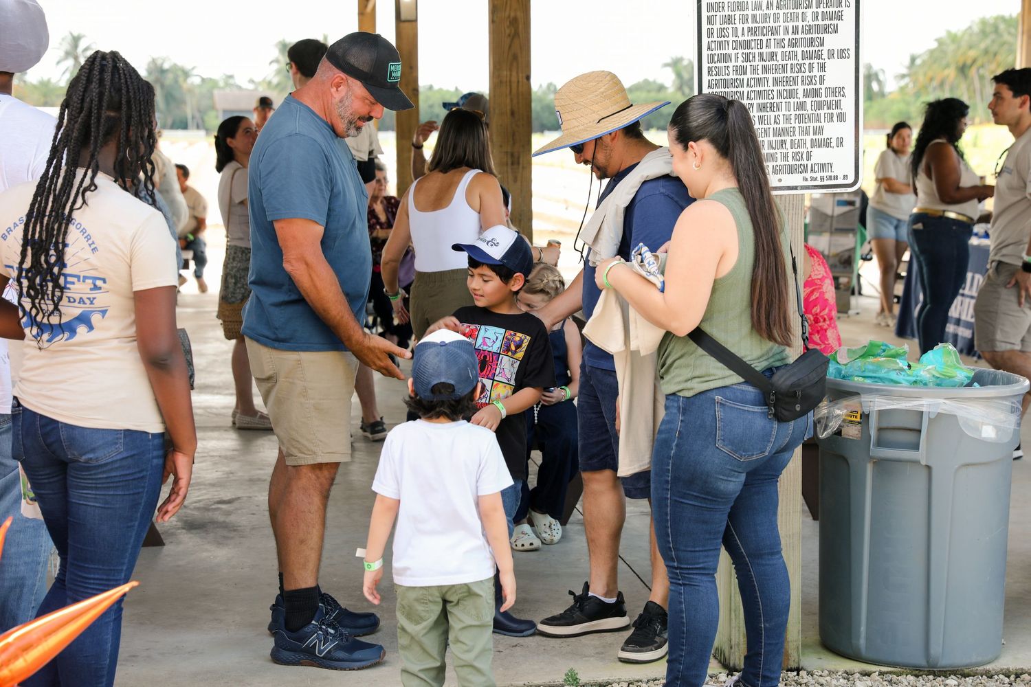 Families with children gather near a sign at an outdoor community event on a sunny day.