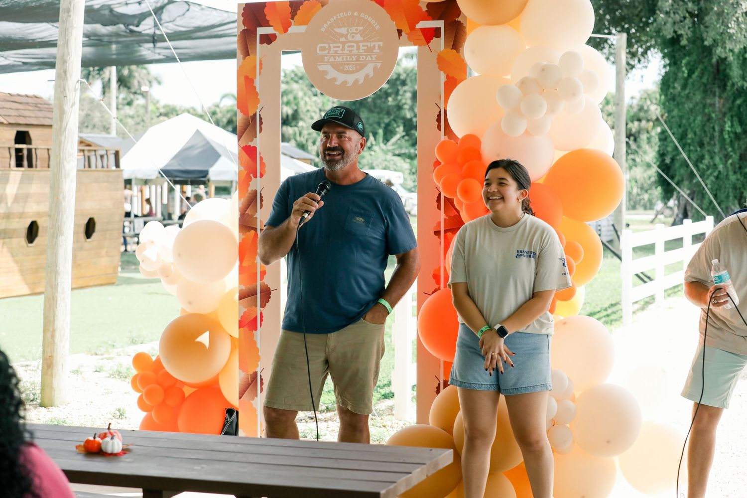 Man speaking at outdoor event under orange and white balloon arch with woman standing beside him.