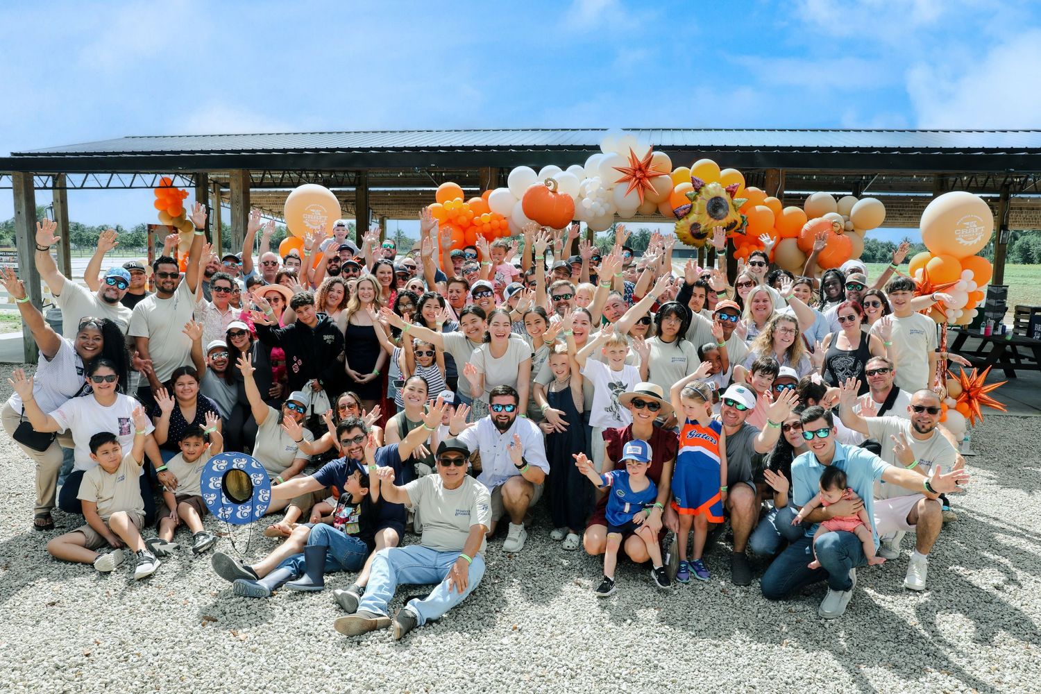 Large group of people celebrating outdoors under a pavilion decorated with orange and white balloons.