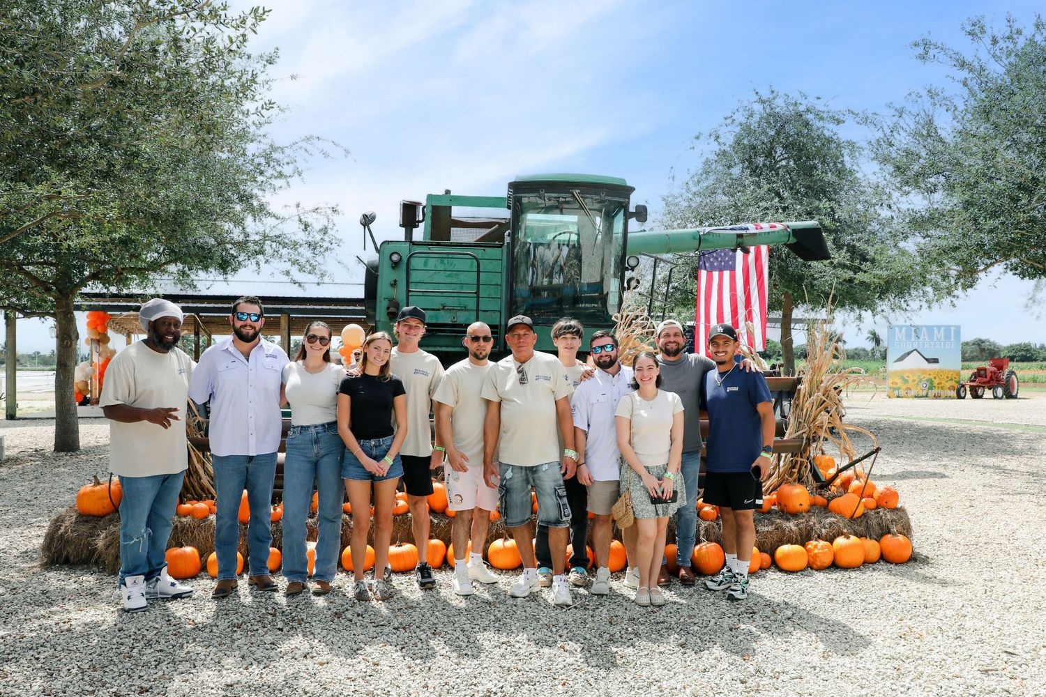 Group of people posing with pumpkins in front of a green combine harvester decorated with an American flag on a sunny day.