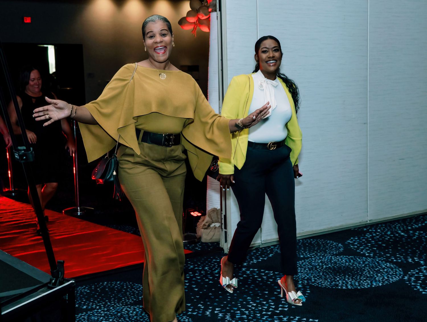 Two stylish women posing and smiling at a formal event, wearing yellow and olive green outfits.